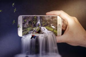 Eine Hand hält ein Smartphone im Kameramodus. Der auf dem Bildschirm abgebildete Wasserfall fließt über das Smartphone hinaus.