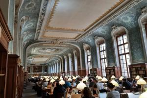 Innenansicht der prächtigen Bibliothek der Sorbonne mit lernenden Studierenden an Tischen. Decken und Wände mit Stuck und Ornamenten, hohe Sprossenfenster, auf den Tischen weiße Leselampen.