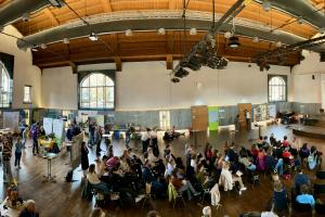 Das Panoramabild zeigt die gut besuchte Lernortmesse in der Mehrzweckhalle der PH Heidelberg, von der Empore aus fotografiert. Links im Bild Messestände, mittig Bestuhlung, rechts die Bühne bei einem der Vorträge.