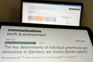 Auf einem Tablet ist der Artikel "The key determinants of individual greenhouse gas emissions are mostly domain-specific" im aktuellen Nature Portfolio Journal Communications Earth & Environment zu sehen. Im Hintergrund sind auf einem Laptop-Screen verschwommen Daten aus der studentischen Untersuchung zu erkennen, die in die Studie eingeflossen sind.