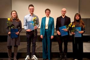 Die Preisträger:innen auf der Bühne mit Prof. Dr. Karin Vach in ihrer Mitte. In den Händen halten sie ihre Urkunde und eine gelbe Rose.