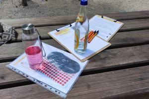Auf einem Holztisch im Freien liegen Arbeitsmaterialien wie ein Klemmbrett, Stifte, eine Tasche etc., außerdem zwei Trinkflaschen.
