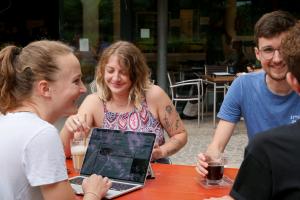 Gutgelaunte Studierende sitzen mit Laptops und Getränken draußen an einem Biertisch