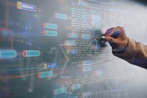 An einer Tafel, die mit blockbasiertem Code überzogen ist, notiert eine Hand mit Kreide mathematische Formeln.