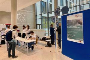Im Foyer des Mathematikons stehen Tische für das Tagungsbüro des Heidelberger Informatiklehrkräftetags, an denen Mitglieder des HSE-Teams die Unterlagen für die neu ankommenden Teilnehmenden ausgebreitet haben. Eine Teilnehmerin nähert sich gerade den Tischen und dem Personal des Tagungsbüros. Rechts im Vordergrund sieht man eine Stellwand, auf der das Plakat des HILT hängt.