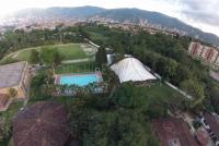 Blick von oben auf das weitläufige grüne Gelände der Deutschen Schule in Medellín mit Kindergartengebäude, Mensa/Kantine, Fußball- und Leichtathletikplatz, Turnhalle, Schwimmbecken und Sportpavillon – im Hintergrund die Stadt, dahinter die Berge | © Tobias Apel