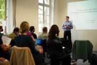 Dr. Florian Grafl beim ersten Cluster-Forum des Forschungs- und Transferclusters „Kulturelles Erbe und Kanonisierung von Wissen“; im Hintergrund die projizierte Präsentation, im Vordergrund Gäste. 