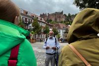 Prof. Dr. Hans Gebhardt auf dem Kornmarkt. Im Hintergrund das Heidelberger Schloss, vorne sind Teilnehmende des Stadtrundgangs zu erkennen.