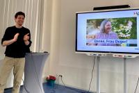 Sebastian Mahner, applaudierend und lachend bei seiner Abschiedsrede; neben ihm ein Smartboard mit Petra Degers Foto und dem Schriftzug „Danke, Frau Deger“.