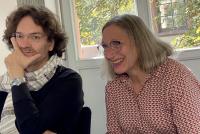 Michael Haus und Petra Deger sitzen nebeneinander am Tisch, beide lächeln. Im Vordergrund Blumen.