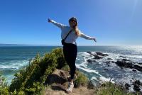 Auf einer Klippe vor dem Meer steht eine junge Frau und reißt glücklich die Arme nach oben.