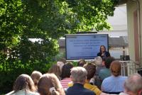 Sitzende Teilnehmende des HSE-Terrassengesprächs sind von hinten zu sehen wie sie nach vorne auf ein Smartboard schauen. Neben dem Smartboard steht Dr. Nicole Aeschbach und begrüßt die Anwesenden zum Thema "BNE in kritischen Zeiten: Perspektiven und Herausforderungen der aktuellen schulischen und hochschulischen Umsetzung". Im Hintergrund ist ein Baum mit voller Blätterpracht zu sehen. 
