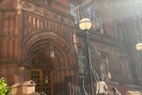 Außenaufnahme eines historischen Campusgebäudes mit steinernem Torbogen-Eingang und Treppe; zwei blaue "Teachers College"-Fahnen hängen an Masten, starkes Sonnenlicht von rechts.