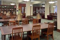 Innenansicht eines Bibliotheks-Lesesaals mit großen Holztischen, Stühlen und Tischlampen; im Hintergrund Bücherregale und Rundbögen.