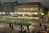 Weitwinkelaufnahme des Teachers-College-Campus bei Nacht; ein hell erleuchtetes Gebäude, Rasenflächen, Wege, Menschen und abgestellte Fahrräder