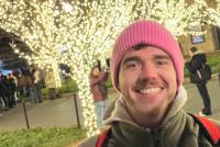 Stipendiat Julius lächelt vor mit Lichtern geschmückten Bäumen; im Hintergrund ein Gebäude und Menschen auf dem Campus des Teachers College bei Nacht.