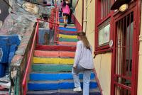 Eine junge Frau geht in der Altstadt von Valparaíso eine Steintreppe mit Stufen in Regenbogenfarben hoch.