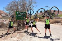 Vier lachende junge Frauen in Fahrradoutfits mit Helmen strecken in der chilenischen Atacama-Wüste ihre Fahrräder über ihren Kopf.