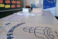 Das Bild zeigt den Tagungstisch mit PH-Jutebeuteln, Programm und Namensschildern. Links im Bild eine Pinnwand mit bunten Karten, rechts das blaue Erasmus+-Plakat.