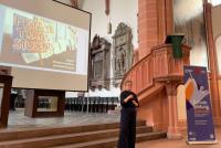 Prof. Dr. Theresa Heyd bei ihremVortrag in der Akademischen Mittagspause am Pult der Peterskirche. Hinter ihr die Schlussfolie des Vortrags mit einem Foto, das ein Demo-Schild mit dem Schriftzug "Protect Trans Students" zeigt. Neben ihr das Roll-up zur Akademischen Mittagspause.