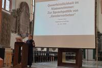 Theresa Heyd am Pult der Peterskirche. Hinter ihr die Startfolie mit dem Vortragstitel "Queerfeindlichkeit im Klassenzimmer: Zur Sprachpolitik von "Genderverboten"
