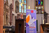 Wiebke Steinleitner bei ihrem Vortrag zur in der Peterskirche. Im Hintergrund das Roll-up zur Akademischen Mittagspause und die Buntglasfenster der Kirche..