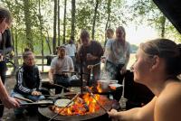 Junge Menschen sitzen gemeinsam an einem Lagerfeuer im finnischen Wald und grillen.